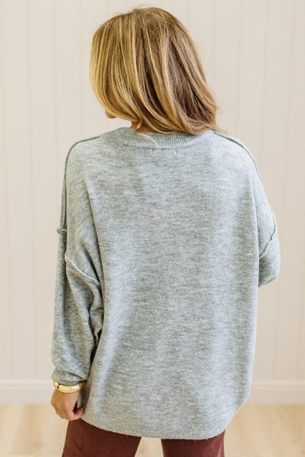 A woman with long blonde hair wearing an oversized light heather gray crewneck sweatshirt and blue jeans, standing against a light beige paneled wall.
