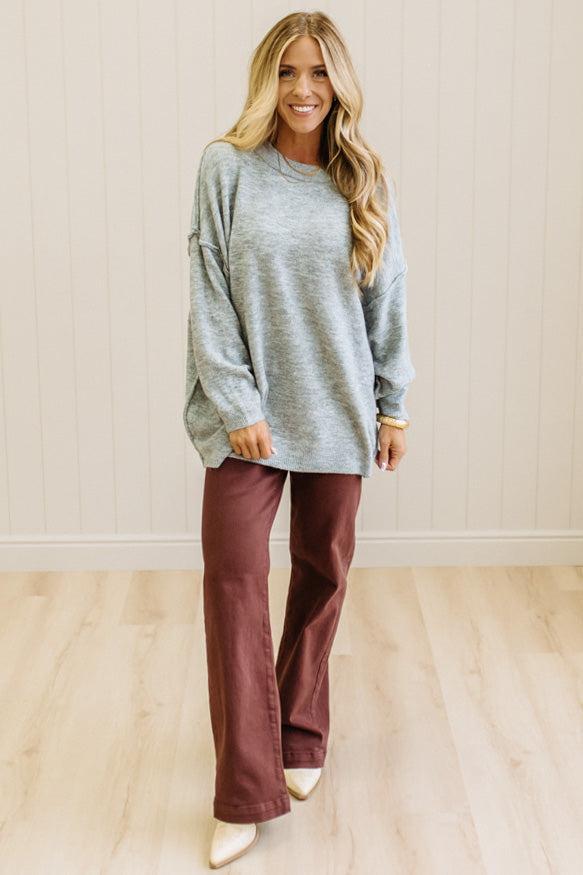 A woman with long blonde hair wearing an oversized light heather gray crewneck sweatshirt and blue jeans, standing against a light beige paneled wall.