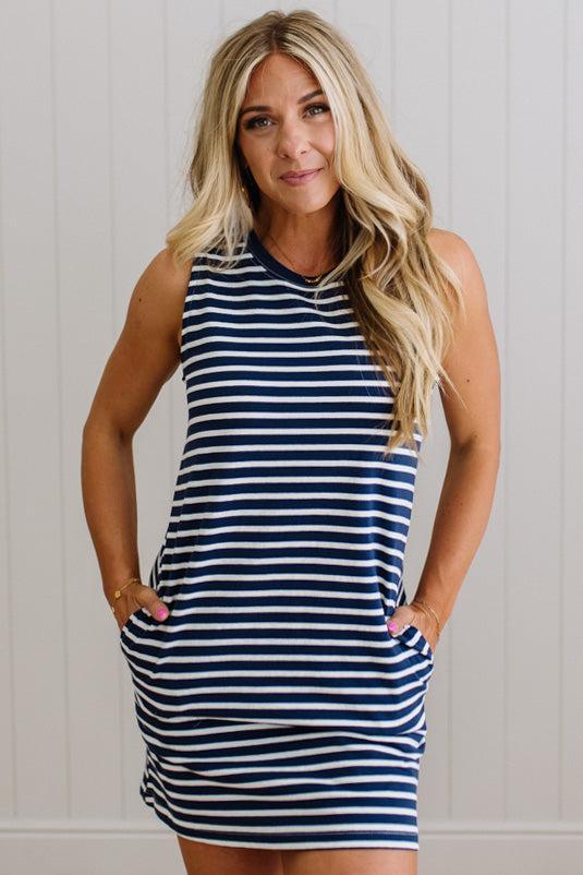 Blonde woman wearing a navy blue and white horizontal striped tank top knee length dress with sandals, standing on a wood floor with neutural backdrop.