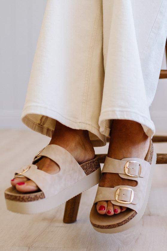 Beige double-strap platform slide sandals with gold-tone buckle details and a cork midsole, shown on a model wearing off-white wide-leg jeans.