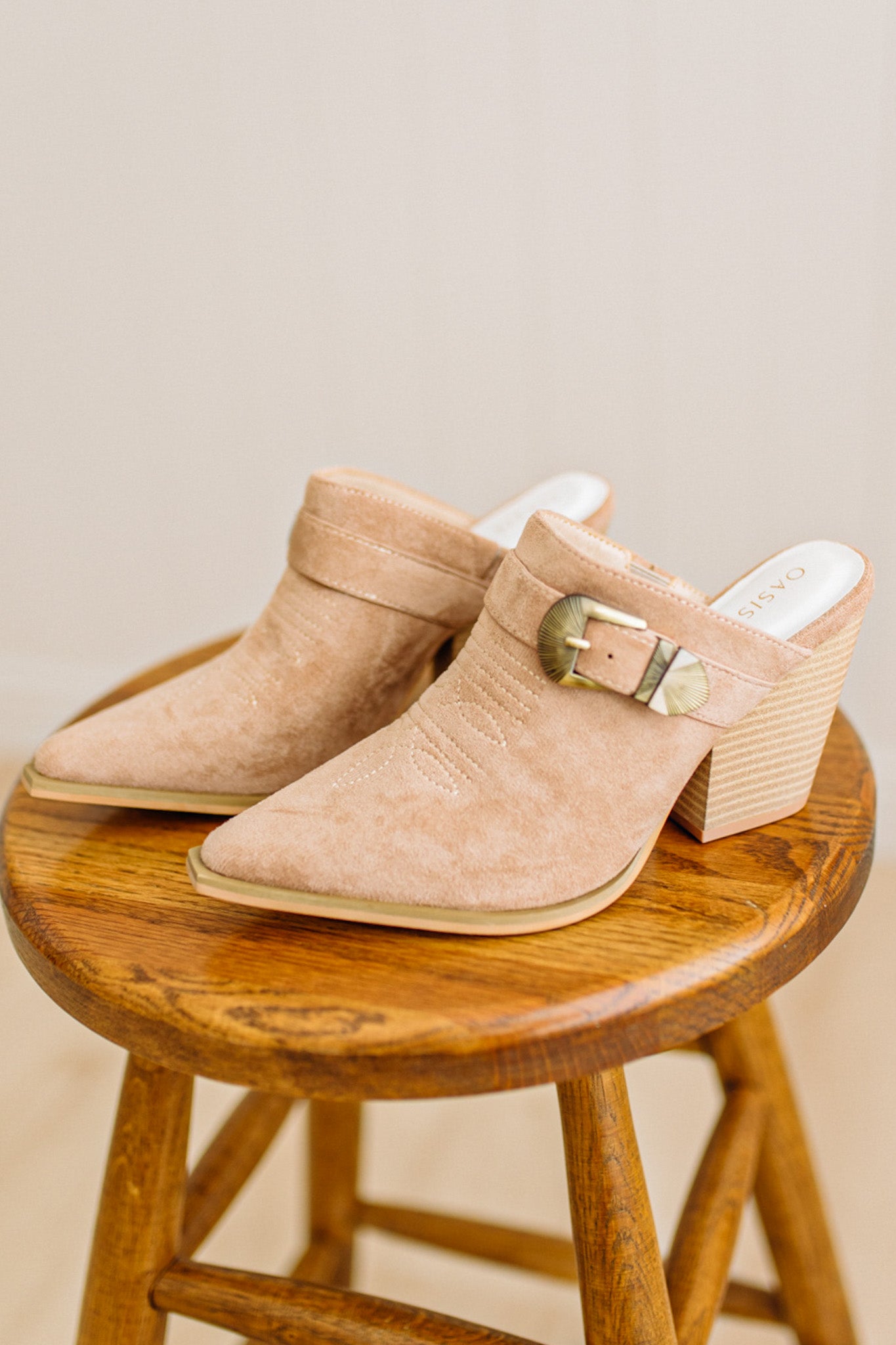 Beige suede mules with wooden block heels on a wooden stool against a beige background