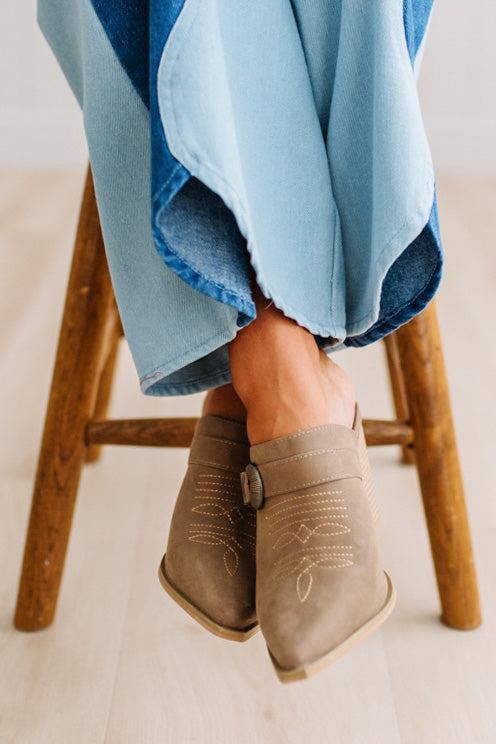 A close-up of a person's feet wearing taupe suede mule boots with decorative stitching and buckle straps, paired with denim two tone jeans on a light wood floor.