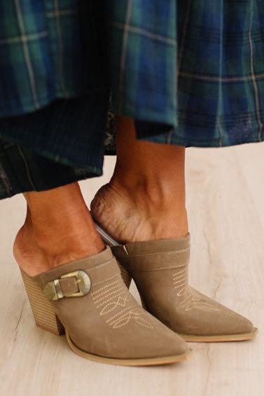 A close-up of a person wearing taupe suede pointed-toe mule boots with a stacked block heel, decorative stitching, and a gold buckle detail, paired with a blue plaid skirt on a light wood floor.