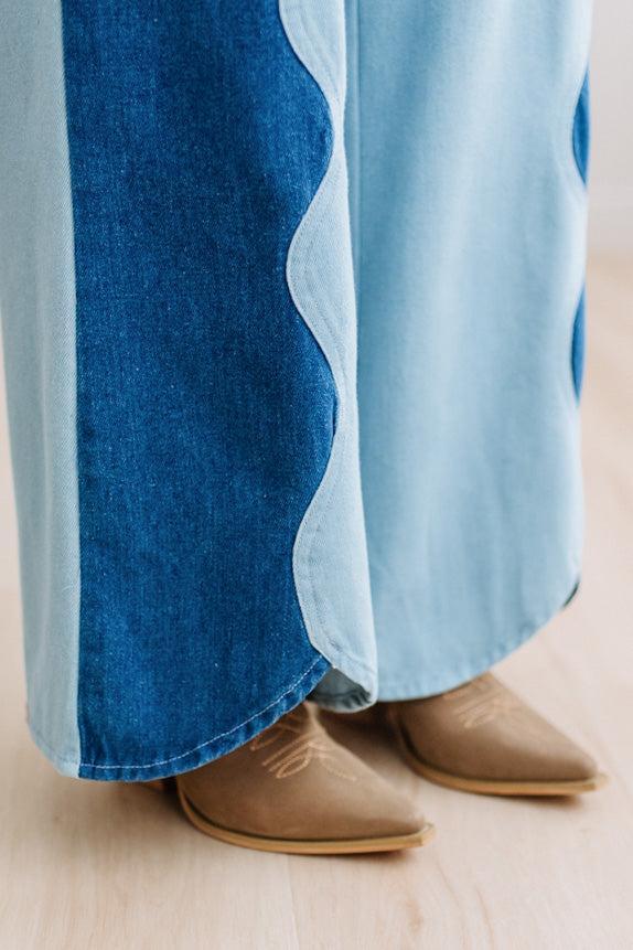A close-up of a person's feet wearing taupe suede mule boots with decorative stitching and buckle straps, paired with denim two tone jeans on a light wood floor.