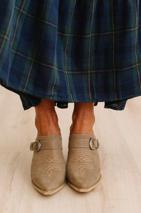 A close-up of a person's feet wearing taupe suede mule boots with decorative stitching and buckle straps, paired with a blue plaid skirt on a light wood floor.
