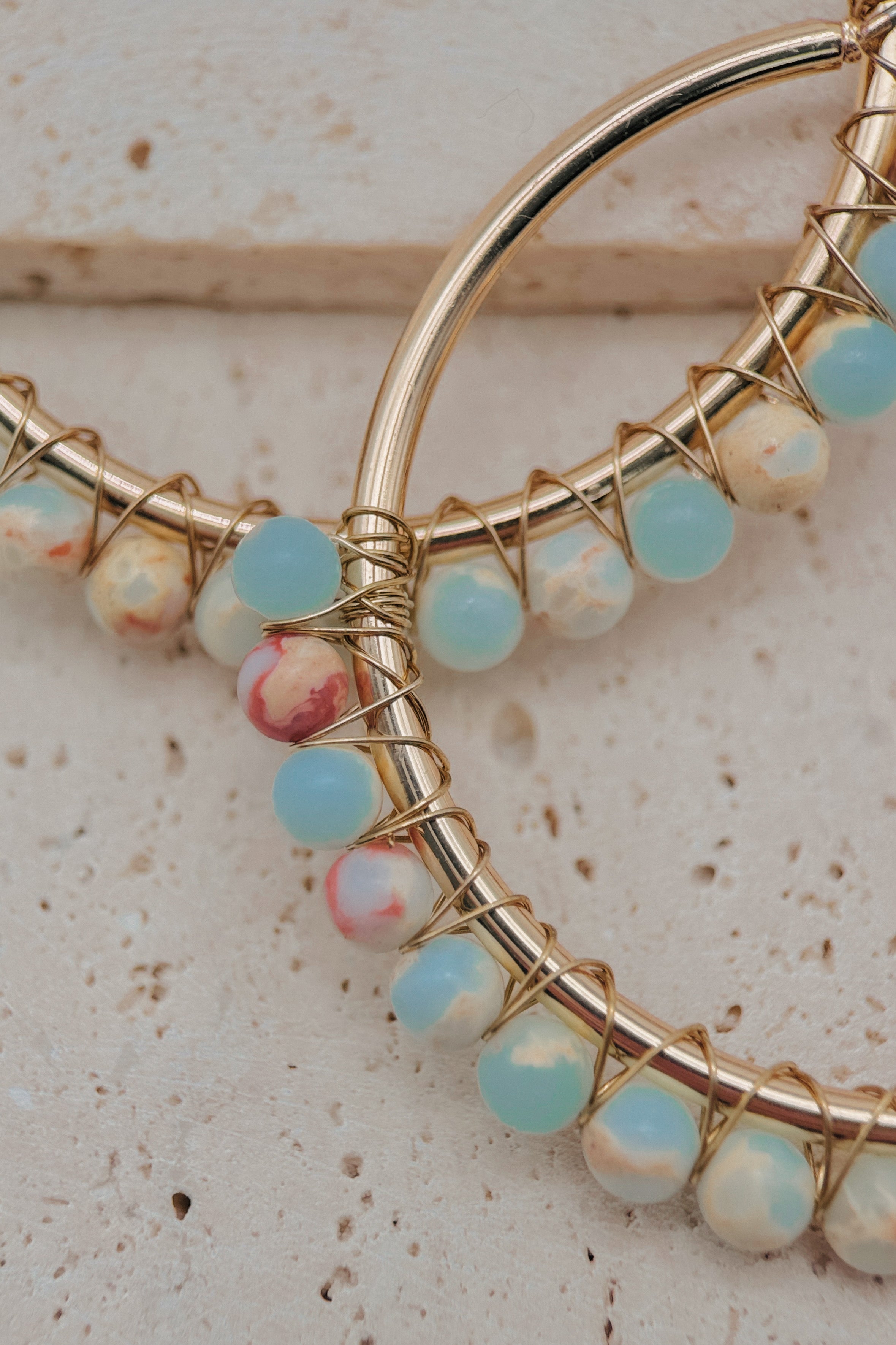 Gold hoop earrings with wrapped light blue and marbled white beads, displayed on a textured beige stone surface.