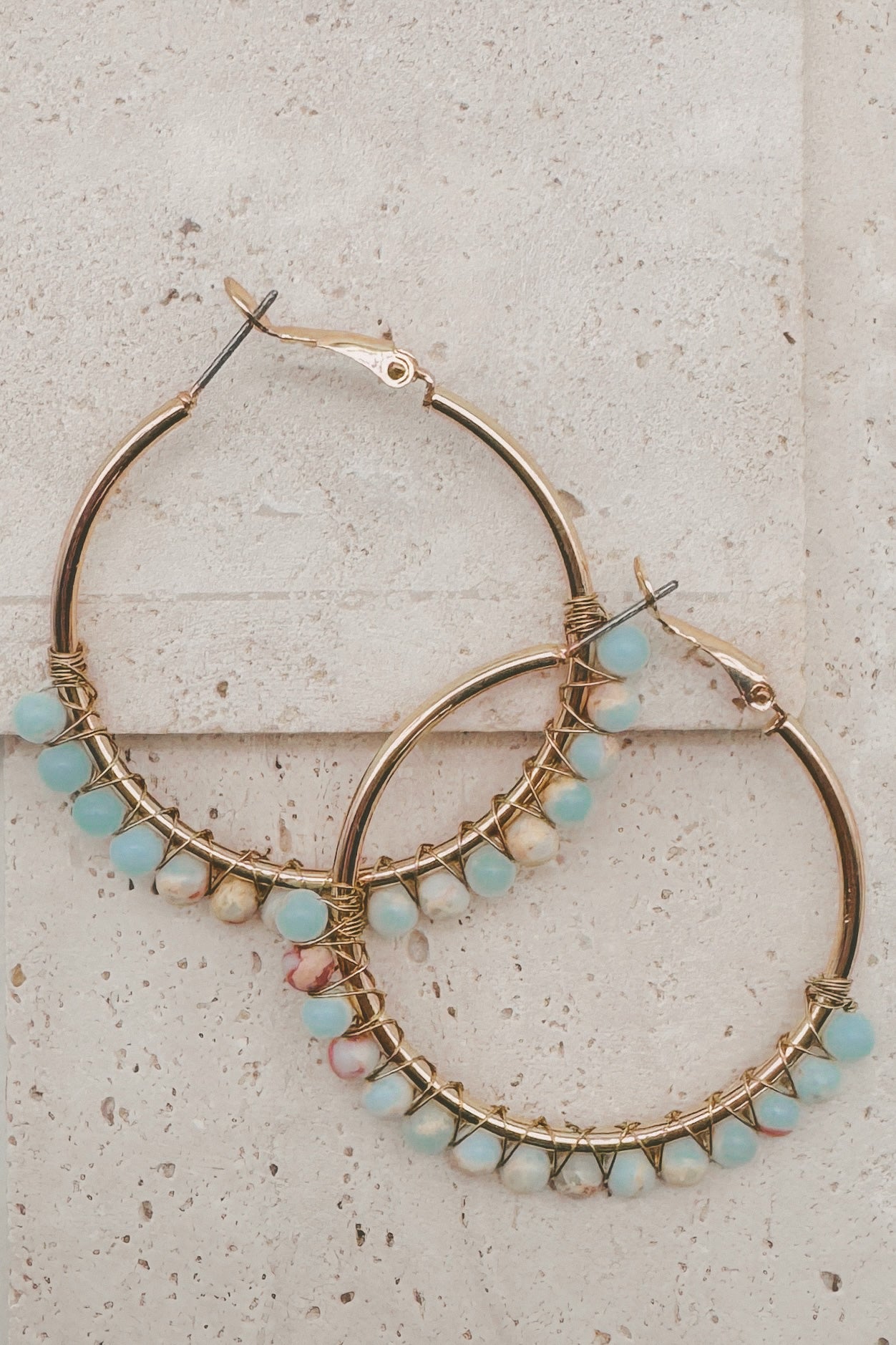 Gold hoop earrings adorned with small light blue and pink beads wrapped in wire, displayed on a light beige stone surface.