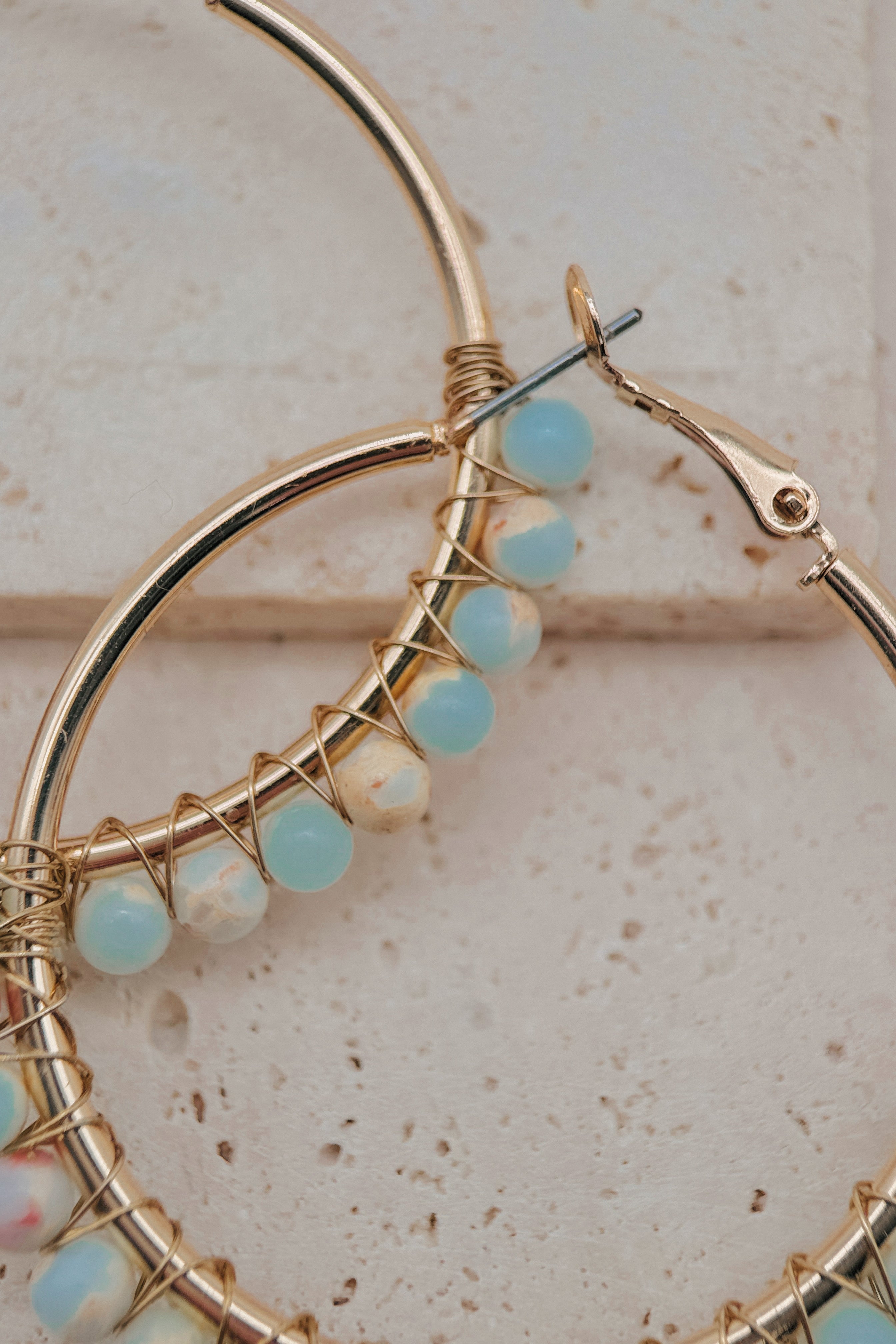 Gold hoop earrings with delicate wire wrapping and small light blue and white beads, shown in a close-up on a textured beige surface.