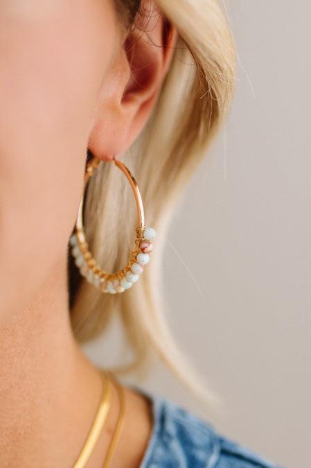 A close-up of a woman wearing a large gold hoop earring adorned with small white pearls and a single pale pink bead, paired with a gold chain necklace.