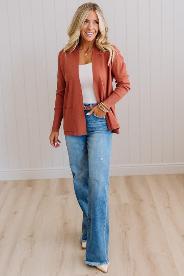 A woman modeling a rust-colored open-front blazer over a white top and blue jeans, standing in a room with light wood floors and white paneled walls.