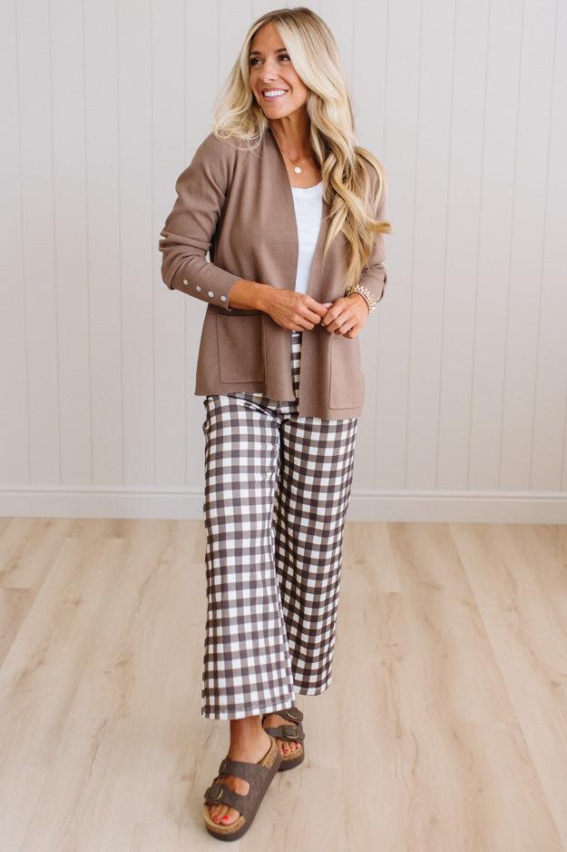 A woman modeling a taupe brown cardigan, brown and white gingham wide-leg pants, a white top, and brown slide sandals in a bright room with light wood floors and white paneled walls.