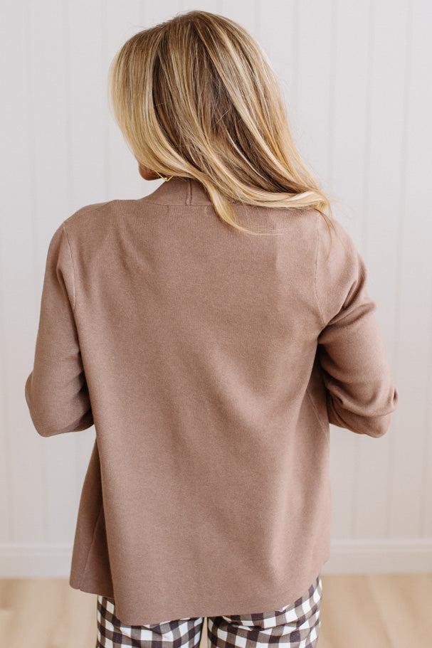 A woman viewed from behind wearing a loose, long-sleeve taupe-brown knit top and brown-and-white checkered pajama pants against a white paneled wall.