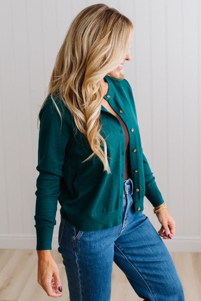 Blonde woman in dark jeans modeling in bright studio wearing a solid green cardigan with brown button front.