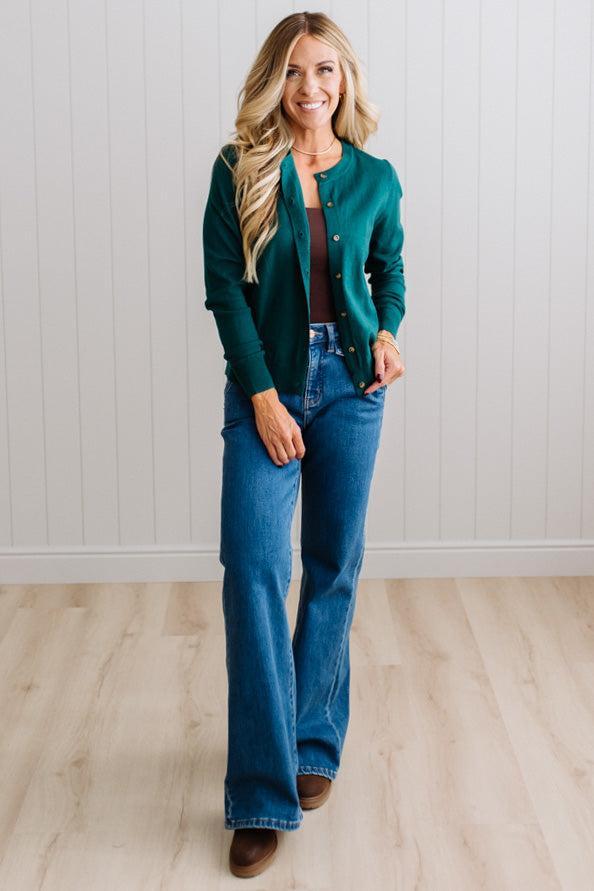 Blonde woman in dark jeans modeling in bright studio wearing a solid green cardigan with brown button front.