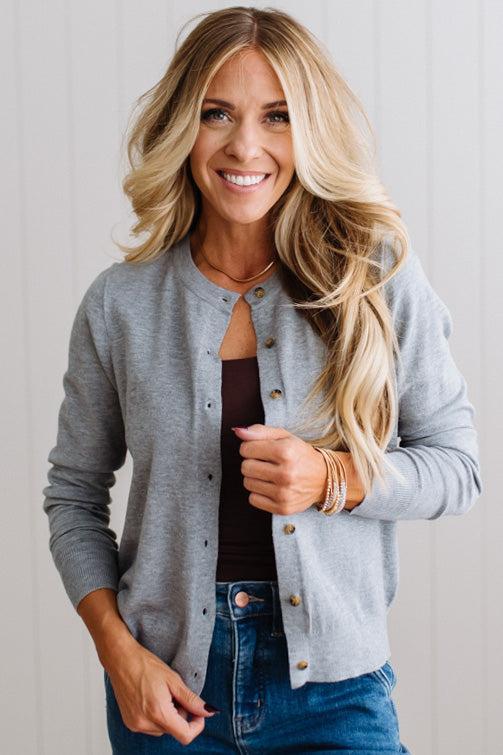 Blonde woman in dark jeans modeling in bright studio wearing a heather grey cardigan with brown button front.