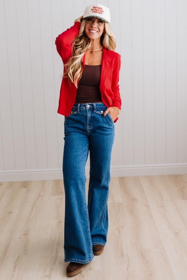 Blonde woman in dark jeans modeling in bright studio wearing a red cardigan with brown button front.
