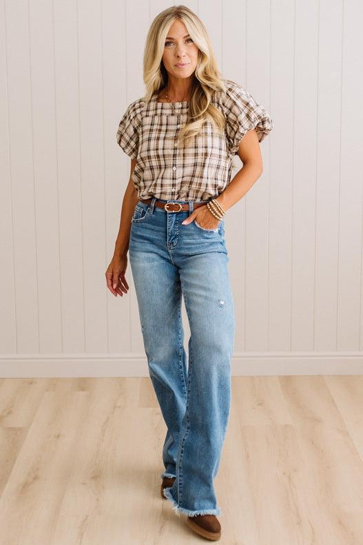 A woman modeling a brown and beige plaid short-sleeve button-up shirt with blue high-waisted flare jeans and brown boots, standing against a white paneled wall and light wood floor.