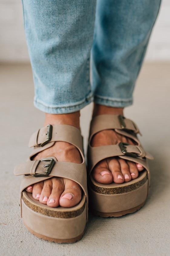 Beige sandals with buckles worn with light blue jeans on a neutral background