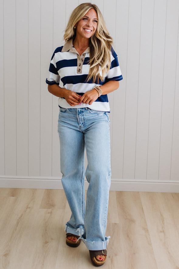 Woman wearing a striped shirt and jeans standing against a white paneled wall.