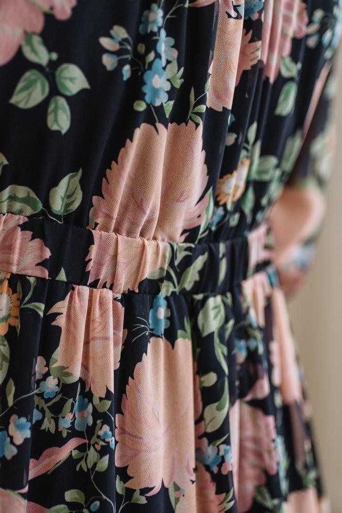 Close-up of a floral dress with a black background, large peach-pink flowers, green leaves, and small blue blossoms. The fabric has a gathered waistband and soft folds.