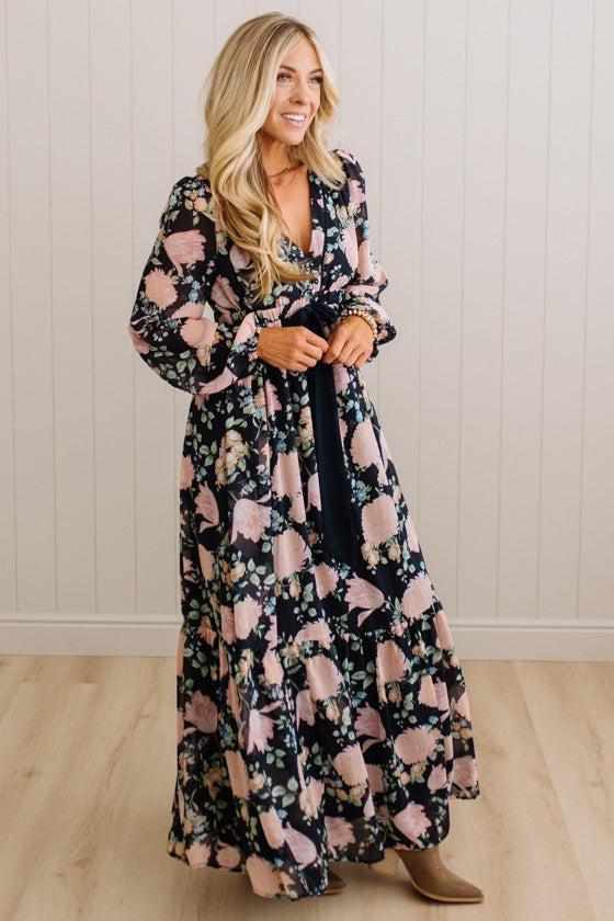 A woman modeling a long-sleeve maxi dress with a black base and a pink floral pattern, standing in front of a white paneled wall and light wood floor.