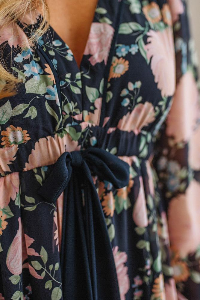 Close-up of a floral wrap dress with a navy base, pink and peach flowers, green leaves, and a matching navy tie belt at the waist.