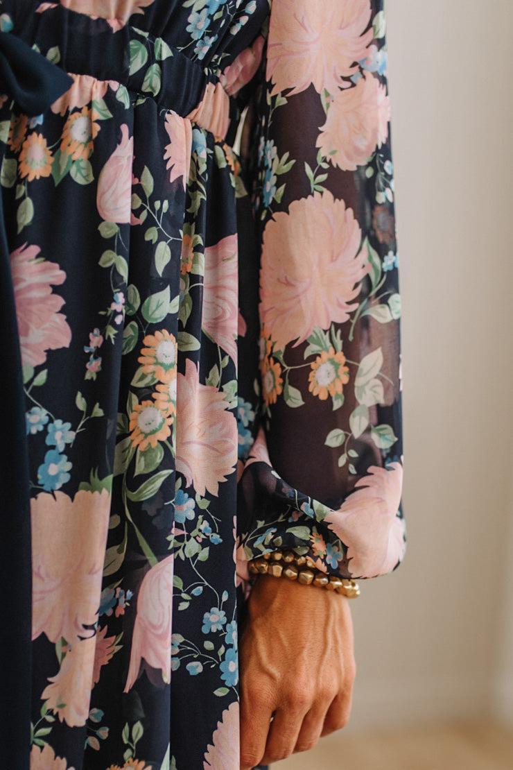 Close-up of a floral long-sleeve dress with a sheer black base and large pink, peach, blue, and green flowers, shown on a model with a beaded bracelet on the wrist.