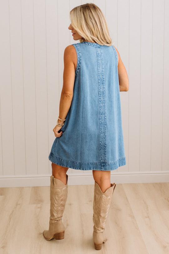 A woman shown from behind wearing a sleeveless light blue denim dress and beige cowboy boots, standing in a room with light wood floors and white paneled walls.