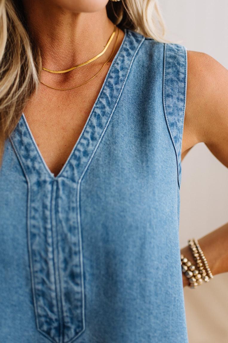 Close-up of a woman wearing a sleeveless light blue denim top with a V-neck and decorative button detail on the front, styled with layered gold necklaces and bracelets.