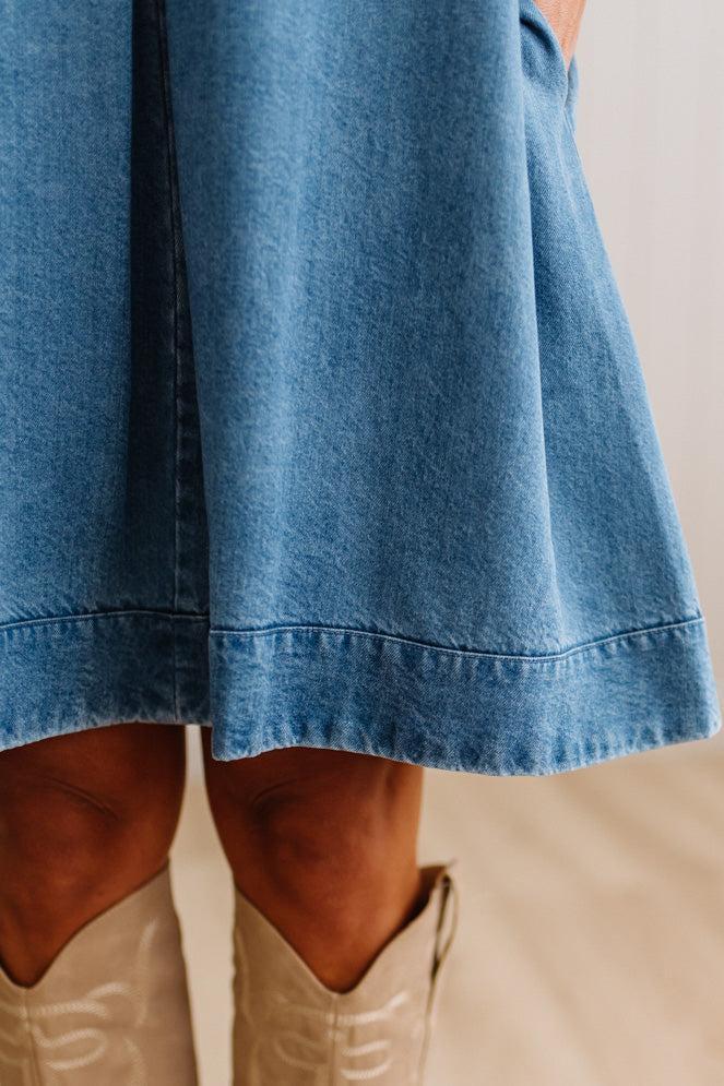 Close-up of a person wearing a blue denim skirt or dress with a wide hem, paired with light beige cowboy boots against a neutral indoor background.