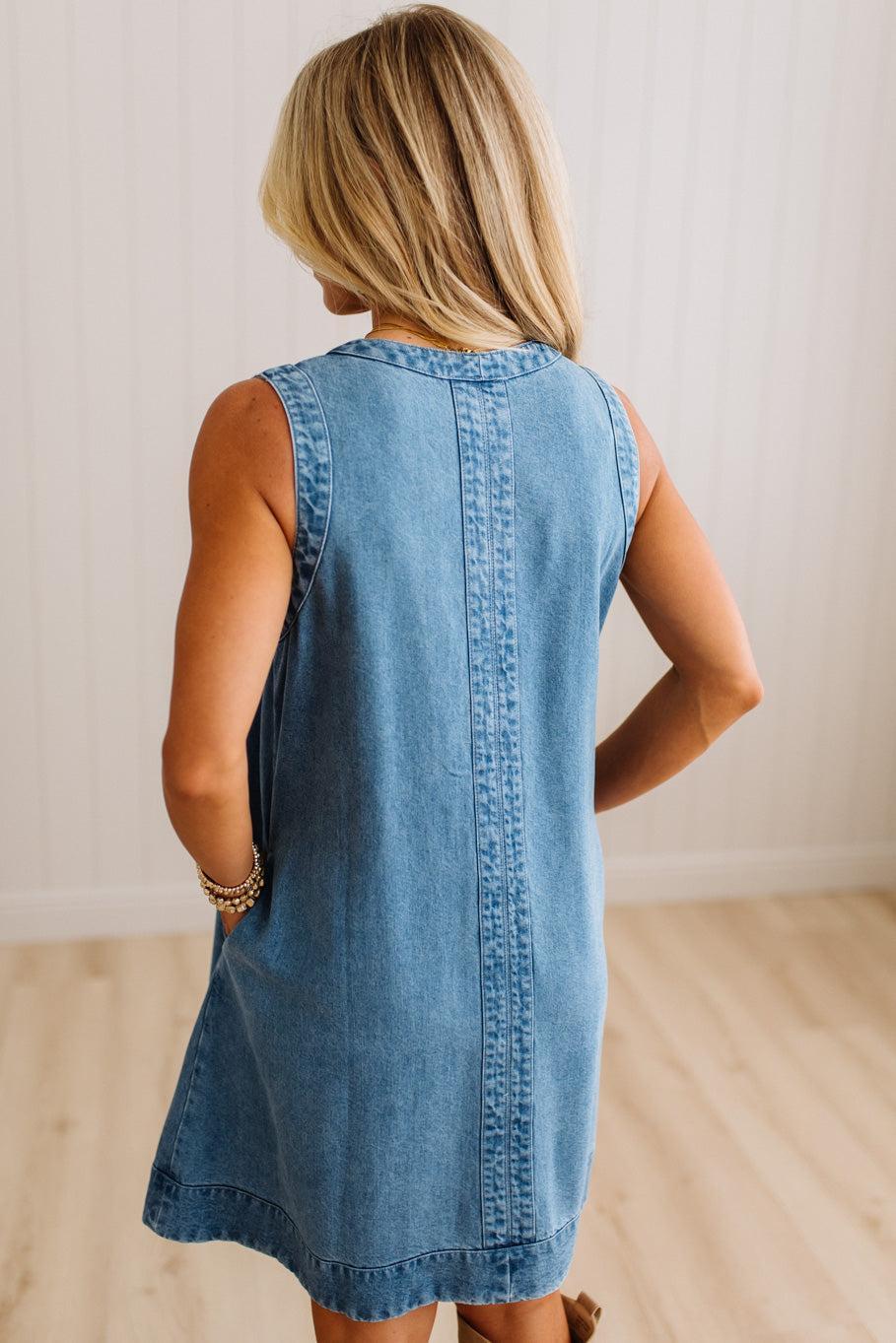 A woman viewed from behind wearing a sleeveless light blue denim dress in a bright indoor room with white paneled walls and light wood flooring.