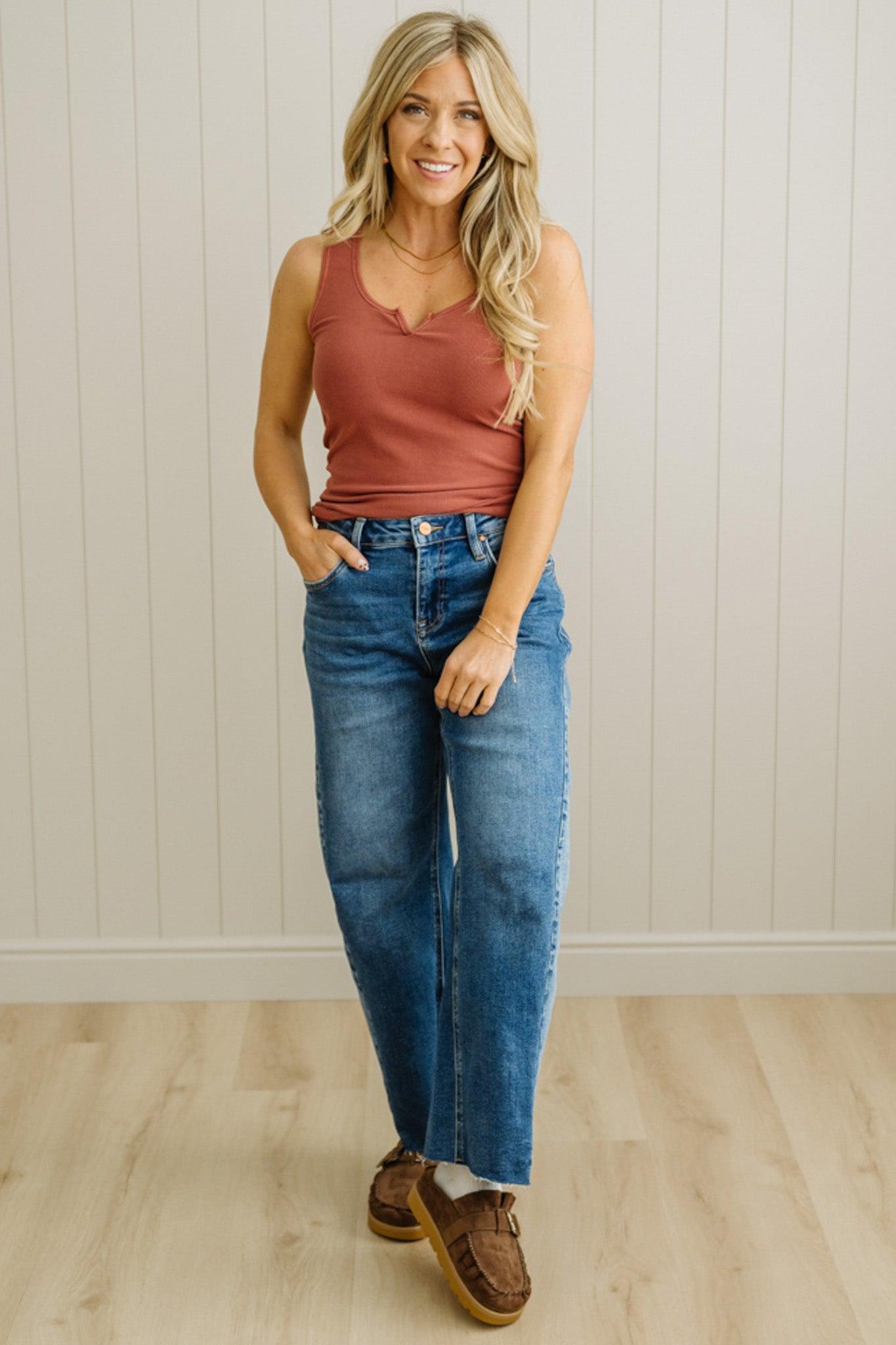Woman wearing a rust tank top and blue jeans standing against a light wooden paneled wall.