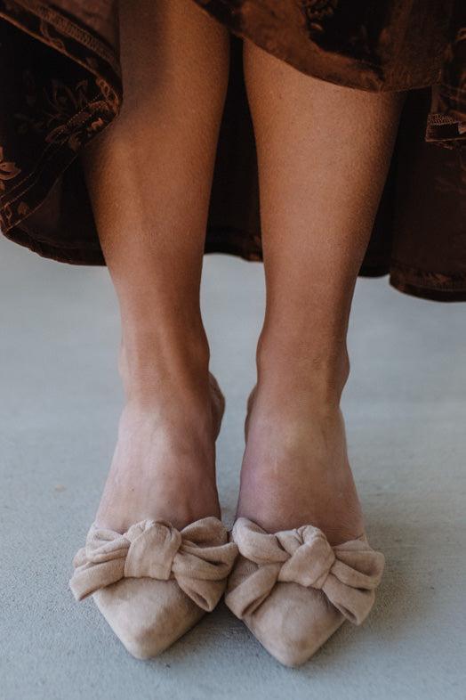 Beige shoes with bow details worn by a person sitting on a brown surface.