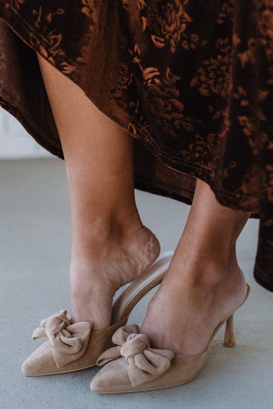 Beige high-heeled shoes with bow details worn by a person in a brown patterned dress.