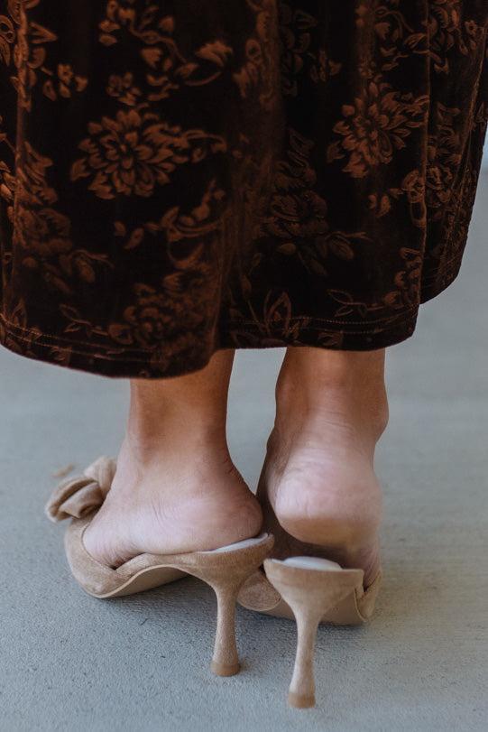 Close-up of feet wearing beige high-heeled shoes with a patterned brown skirt.