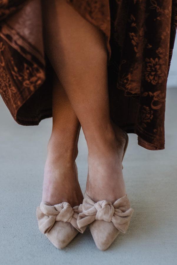 Beige high-heeled shoes with bow details worn by a person in a brown patterned garment.