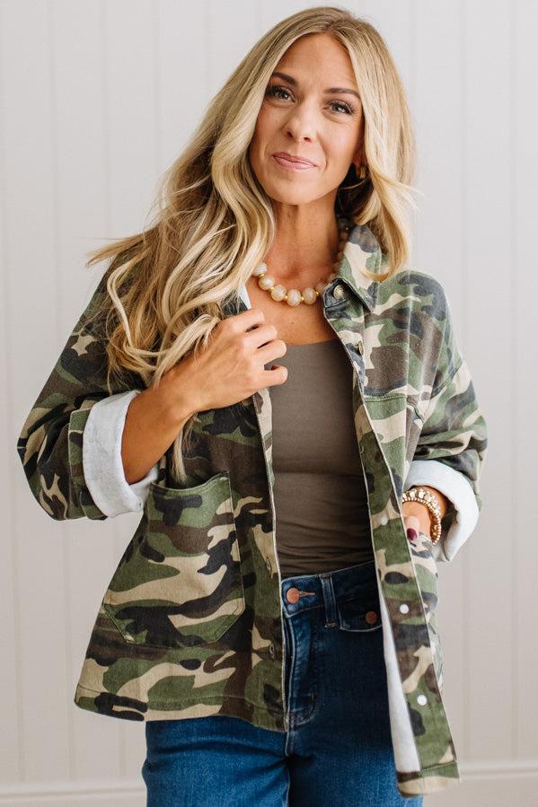 A woman wearing a green and brown camouflage flannel button-up shirt over a taupe tank top and blue jeans, standing against a white paneled wall.