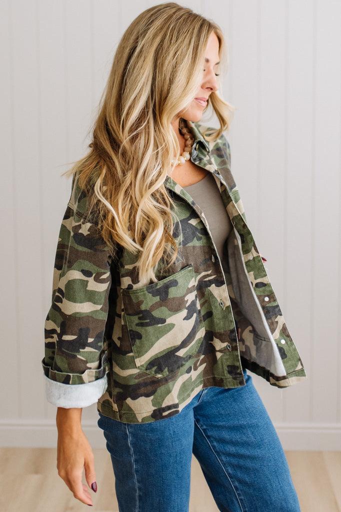 A woman wearing a green and brown camouflage utility jacket over a gray top and blue jeans, standing against a white paneled wall.