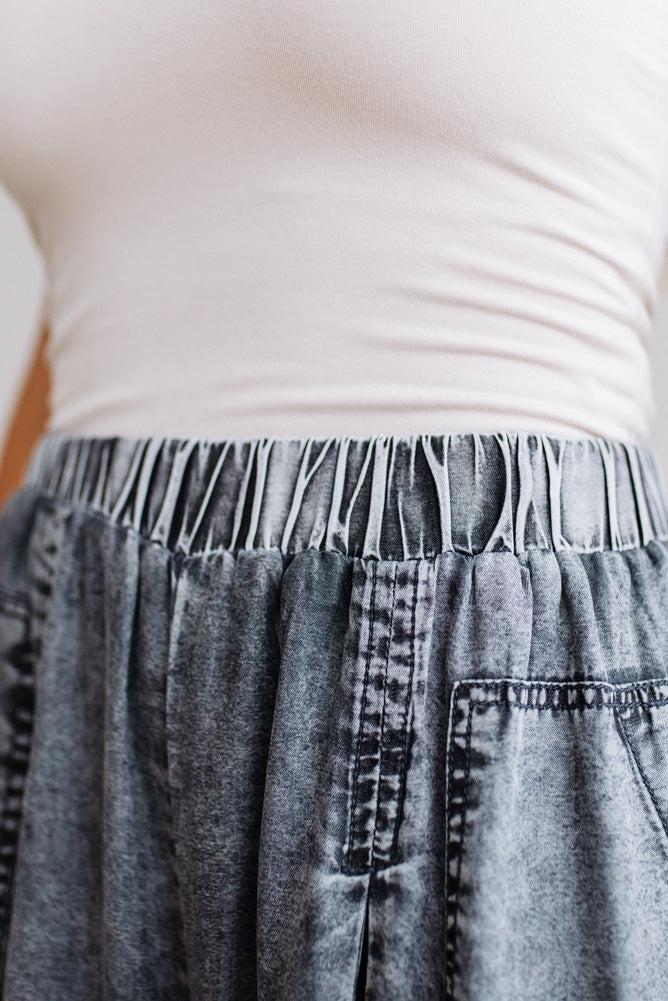 WOMAN IN A BRIGHTLY LIT STUDIO WEARING A TANK WITH CHAMBRAY ELASTIC WAIST POUCH POCKET WIDE LEG CULOTES PANTS IN GREY.