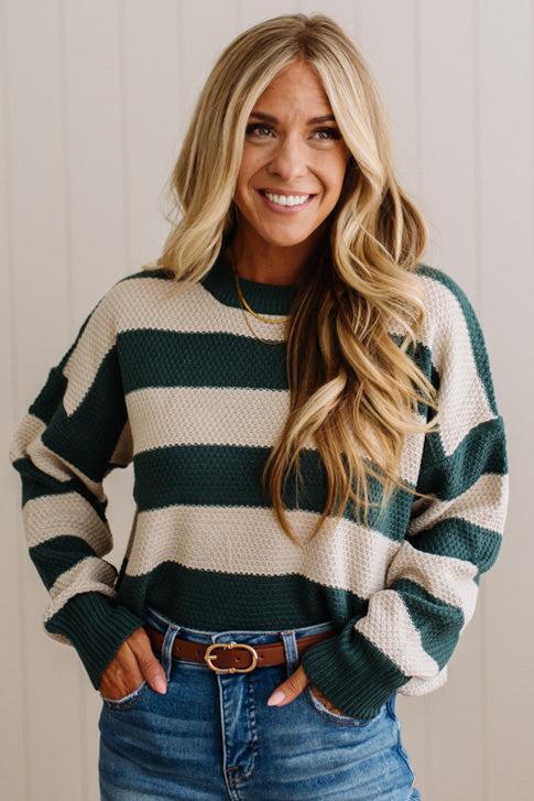 A smiling blonde woman wearing a dark green and cream horizontally striped knit sweater, tucked into blue jeans with a brown belt, standing against a light beige paneled wall.