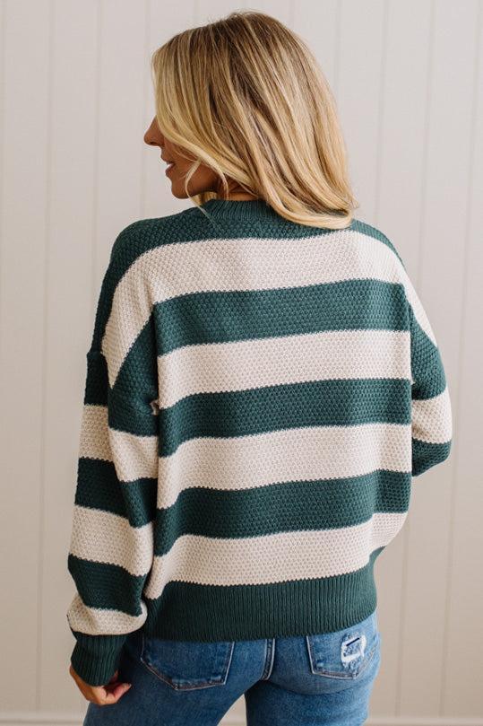 A woman shown from the back wearing a dark green and cream striped knit sweater with blue jeans, set against a light beige paneled wall.