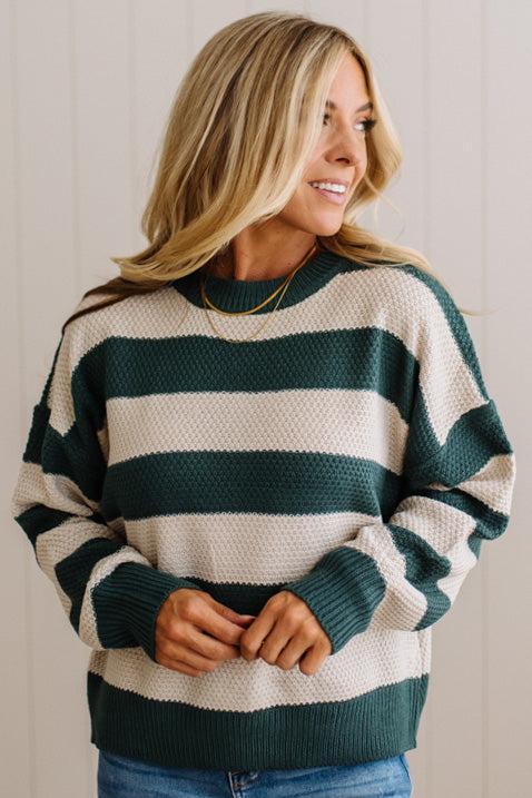A woman wearing a dark green and cream waffle-knit sweater with horizontal stripes, paired with blue jeans and a delicate gold necklace, set against a light beige paneled wall.