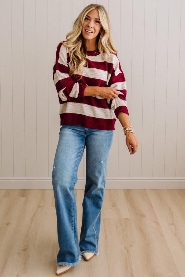 A woman modeling a long-sleeve knit sweater with bold horizontal burgundy and cream stripes, paired with light-wash blue flare jeans and beige pointed-toe heels, standing against a white paneled wall and light wood floor.