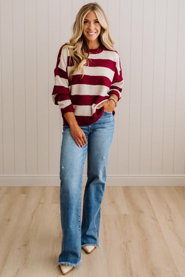 A woman modeling a red and cream striped knit sweater with light-wash blue jeans, standing in front of a white paneled wall and light wood floor.