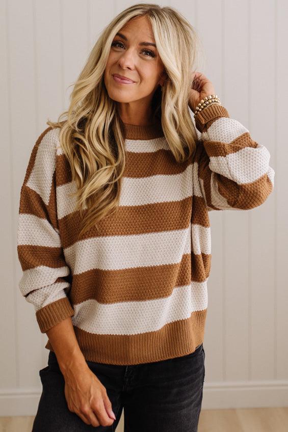 A woman wearing a brown and cream horizontally striped knit sweater with textured waffle knit sleeves, paired with black jeans, standing against a white paneled wall.