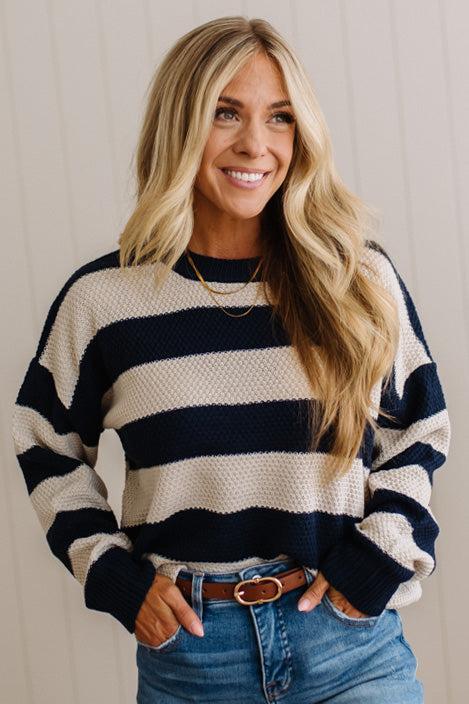 A smiling blonde woman wearing a navy and cream horizontally striped knit sweater, blue jeans, and a brown belt, standing against a light beige paneled wall.