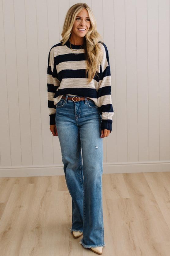 A woman modeling a navy and cream striped long-sleeve knit sweater paired with blue jeans, standing in a room with light wood floors and white paneled walls.