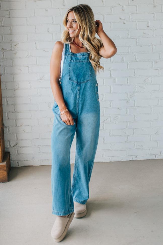 Woman wearing blue denim overalls standing against a white brick wall.