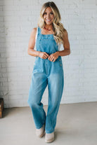 Woman wearing light blue denim overalls standing against a white brick wall.