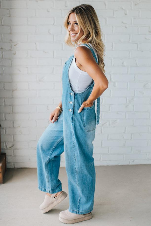 Woman wearing a denim overalls outfit against a white brick wall.